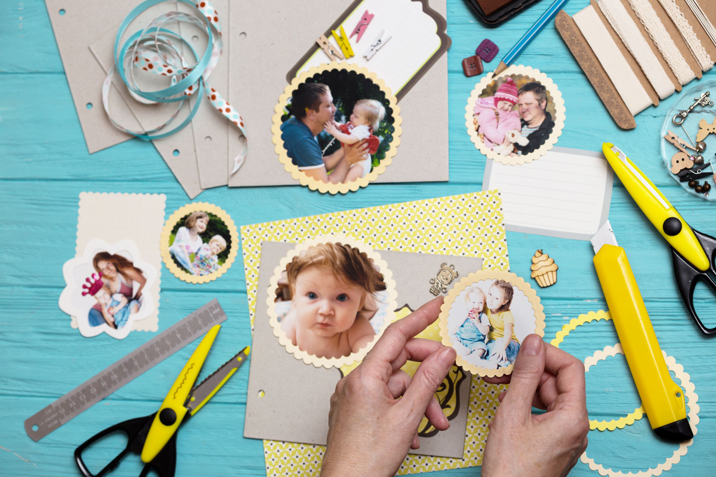Hands holding a scalloped photo cutout of two children, surrounded by scrapbook supplies and other family photo cutouts on a blue wooden surface.