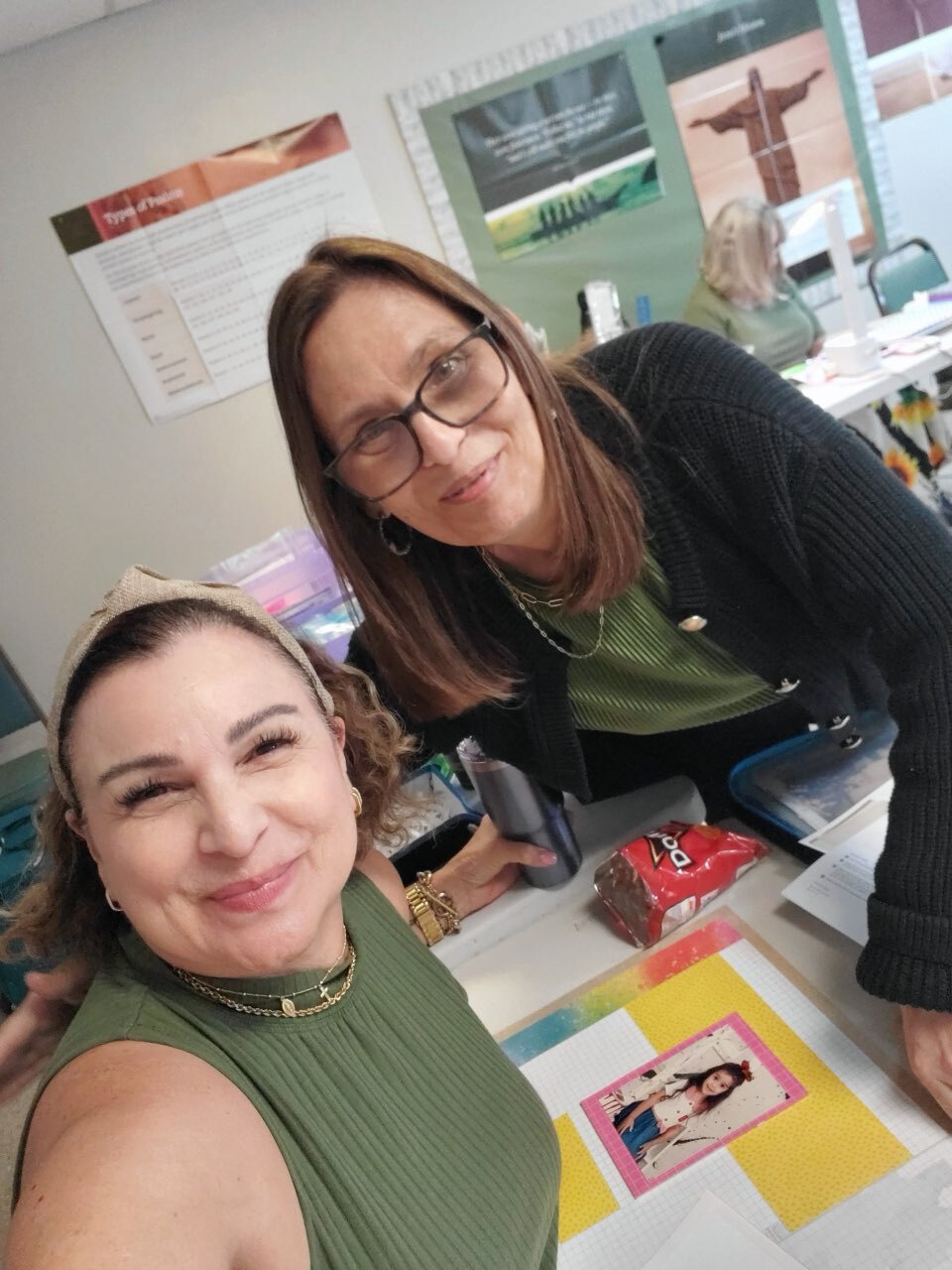 Two smiling women leaning over a table with colorful papers and a photo, in a classroom setting.