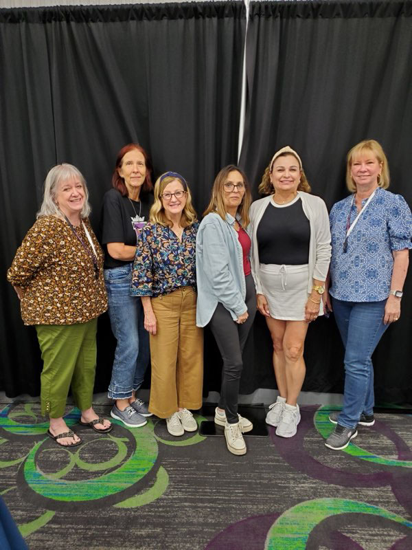 Group of six women standing and smiling in front of black curtains on a patterned carpet.