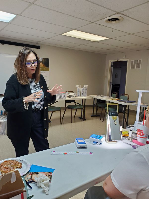 Woman wearing glasses and a black cardigan standing and gesturing with her hands in a room with tables and chairs, facing a person seated at a table with a pizza slice, phone, and large drink cup.