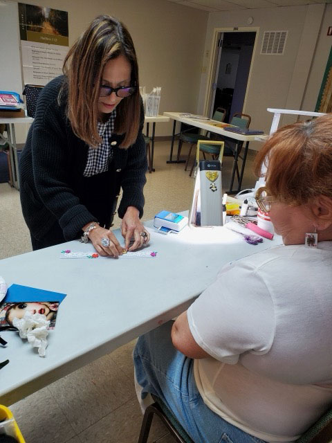 Two women at a table with one woman standing and working on a craft project while the other watches seated.