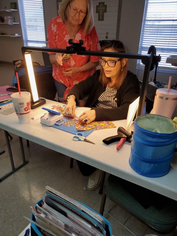 Two women working on a colorful puzzle at a well-lit table with crafting tools and drinks nearby.